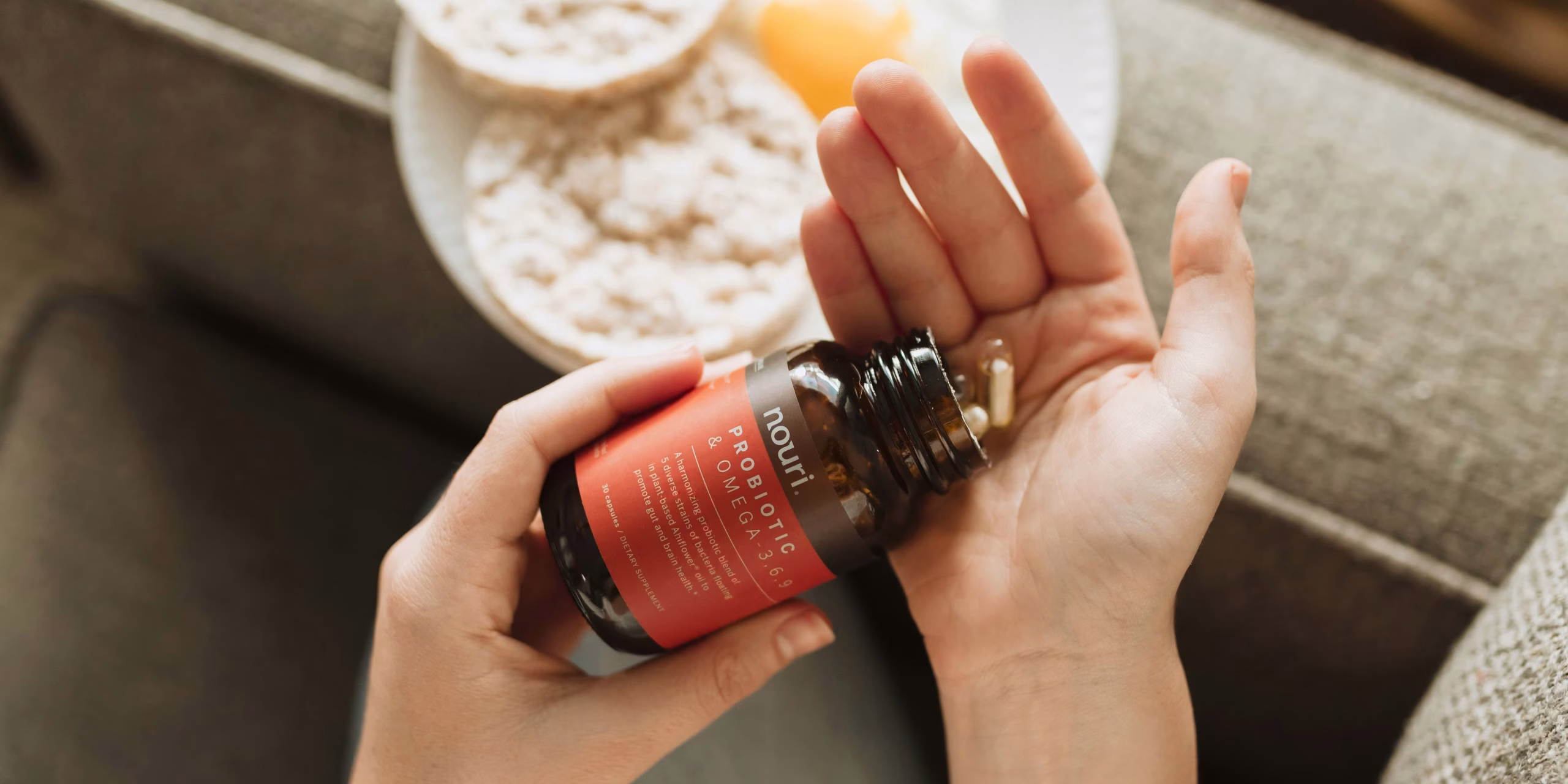 Pessoa colocando na mão comprimidos de omega 3 com um prato no sofá, com o intuito de mostrar o suplemento.